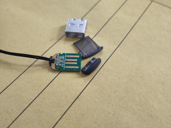 Ein Nitrokey 3 A Mini liegt auf einem Schnellhefter. Die Platine liegt frei, da das Plastik des Gehäuses gebrochen ist und somit das gesamte Gehäuse haltlos ist.