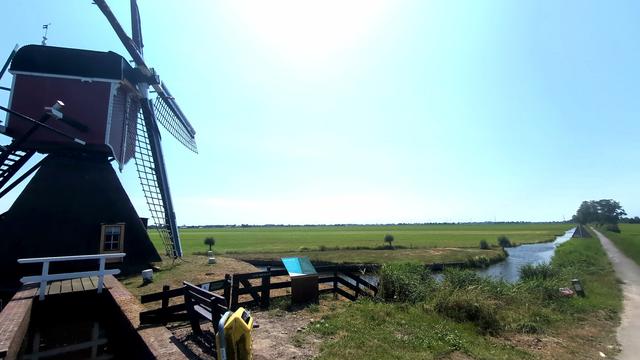 A windmill, behind which you see fields. There is a small road for walking/biking/skating on the left side with a ditvh nect to it seperating it from the fields.