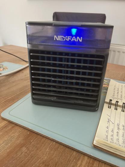 A picture of a dark grey fan based cooler. We can see a little water reservoir with a blue light. Below is the fan outlet with the frozen sponge filet just visible .