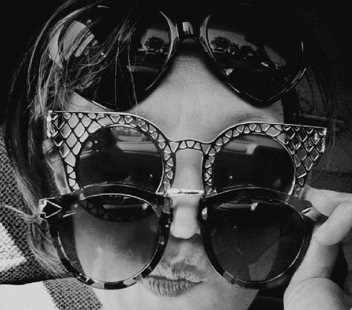 Monochrome, close-up shot. The subject's face fills the frame, strategically layered with three pairs of sunglasses. Dark, heart-shaped sunglasses sit high, reflections mirror a street or landscape. The middle pair feature a detailed, patterned frame. The lowest sun glasses are held with a hand out of view. The face displays a slightly puckered mouth, generating a playful image.