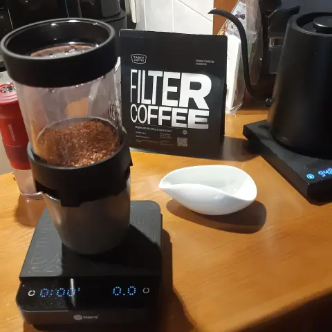 The image shows a coffee brewing setup. In the foreground, there is a coffee maker with a transparent container filled with ground coffee. The container is placed on a digital scale displaying "0.0." Behind it, there is a package labeled "FILTER COFFEE." To the right, there is a small white dish. The background includes other coffee-making equipment, but specific details are not visible.