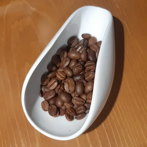The image shows a white, teardrop-shaped dish containing roasted coffee beans. The dish is placed on a wooden surface. The coffee beans are light brown and glossy, filling the dish.