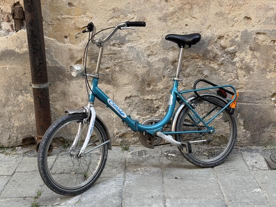 A turquoise bicycle with a unique frame design is leaning against a textured, weathered wall. It features a front light and a rear rack, and the ground is paved with stone tiles.