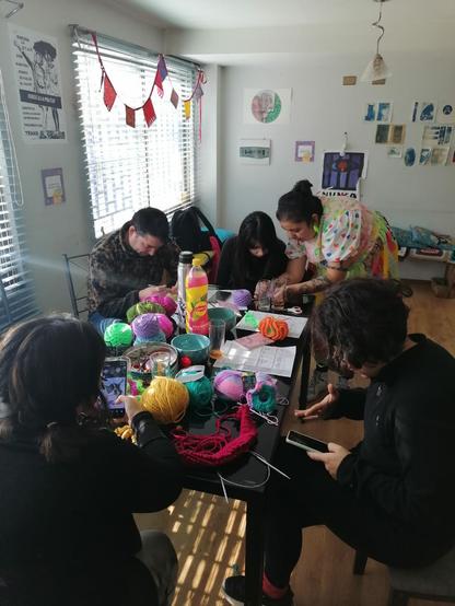 Fotografía del Taller Jaqueo Textil, en la imagen se muestra al rededor de una mesa llena de lanas y herramientas para tejer a un grupo de personas concentradas tejiendo a crochet.
