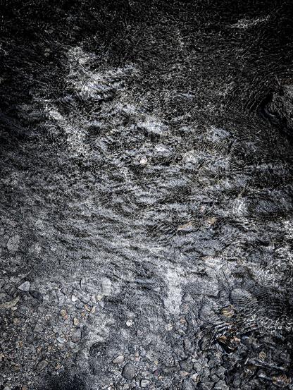 A dark, high-contrast, top-down shot of water flowing over a rocky, uneven surface. The water itself shows ripples, small waves, and streaks of light.