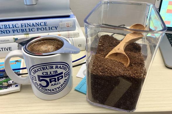 Coffee is brewing in a strainer inside a mug. The mug is white with the slightly oval navy blue logo of the QRP Amateur Radio Club International. The logo prominently features the letters QRP, but the Q is stylized heavily and looks like a five with a long top bar that extends over the R and P. Behind the mug is a stack of books with a monitor on top. Next to the mug is a plastic container of coffee grounds with a wooden measuring spoon inside.