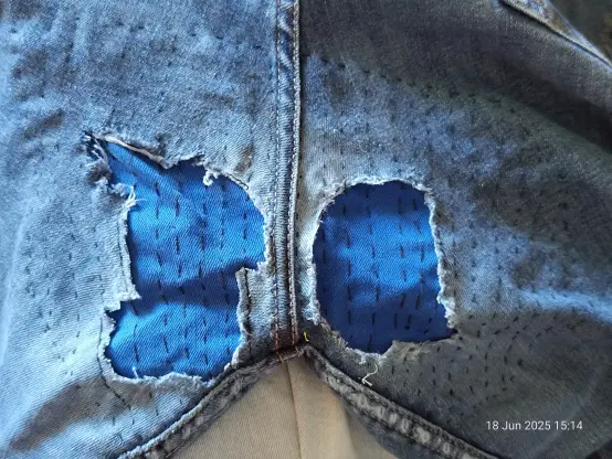 Photo of the seat of a pair of jeans. 2 large holes can be seen on either side of the central buttcrack seam. A bright blue fabric has been used to patch the holes, and the surrounding denim of the jeans is peppered with semi-regular stitches going in up & down in rows parallel to the middle seam. The edges of the holes are quiet freyed in places, even tho I did try to trim them a bit before hand. Dark/medium blue denim with black stitches