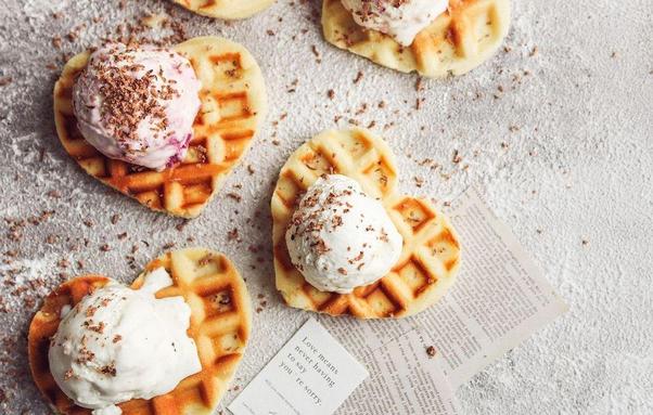 A flatlay of heart-shaped waffles topped with scoops of ice cream, one pink and one white, garnished with chocolate shavings. A few pieces of text are visible in the background, along with a light dusting of powdered sugar.