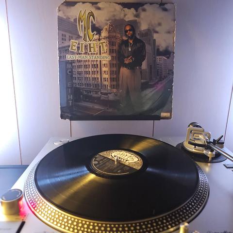 A vinyl record sits on a turntable. Behind the turntable, a vinyl album outer sleeve is displayed. The front cover shows MC Eiht standing in a city street. He's as tall as some of the building behind him.