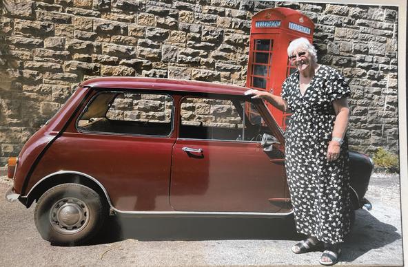 A woman in a long patterned black / grey dress is posing in front of a red Austin Mini. Behind the car is a stone wall with a red telephone box