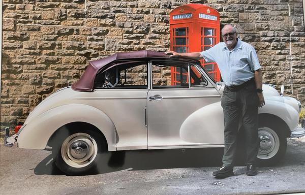 An absolutely stunningly handsome man is posing in front of a white Morris Minor car that has a maroon hood. The man is stylishly displaying a pair of green trousers and a light green short sleeve shirt.
Behind the car is a stone wall with a red telephone box