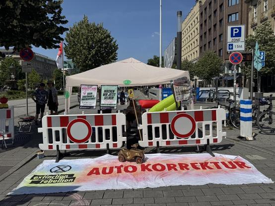 Zu sehen ist der Stand der Kundgebung mit einem großen Banner davor, auf dem AUTOKORREKTUR steht. Daneben das Logo von Extinction Rebellion.