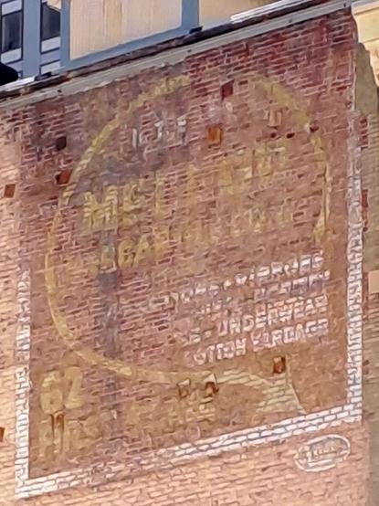 A fading painted sign high up on a brick wall with lettering set inside a circle that curves out bottom right to form an arrow pointing down and to the right.