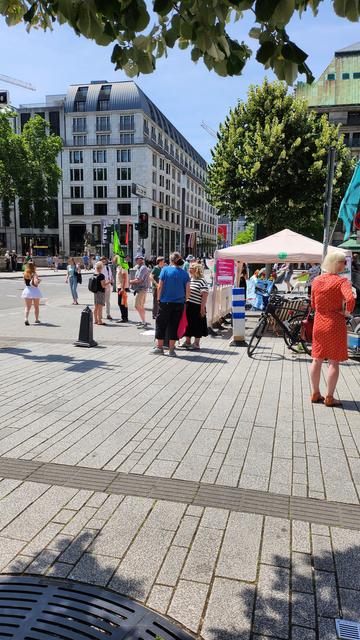Zu sehen ist der Stand der Kundgebung mit diversen Passanten davor. Lukas von den Grünen unterhält sich mit jemandem.