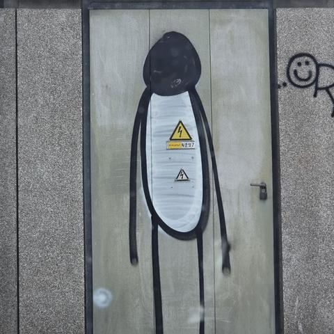 Foto af grafitti tændstiksmand med sort hoved og hvid krop, malet på el-skur, med gul advarselstrekant (ikke overmalet) lige der hvor hjertet ville være.