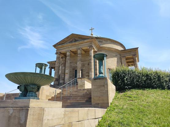 Das vierte Bild zeigt ein klassizistisches Gebäude (die Grabkapelle) auf einem Hügel, das von einer Freitreppe und Säulen geprägt ist. Vor dem Gebäude stehen zwei große, dekorative Bronzeschalen oder Brunnen. Das Gebäude hat eine symmetrische Fassade mit einem Dreiecksgiebel und einem Kreuz auf der Spitze. Der Himmel darüber ist klar und blau, was dem Gebäude ein majestätisches Aussehen verleiht.