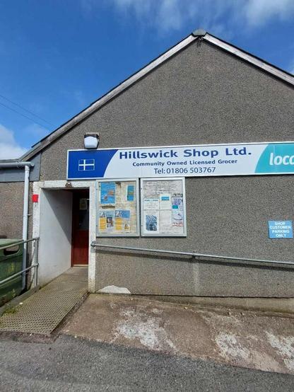 A grey simple building without windows and a single entrance. A sign almost as wide as the wall says 'Hillswick Shop Ltd. Community owned licensed grocer' and their phone number.