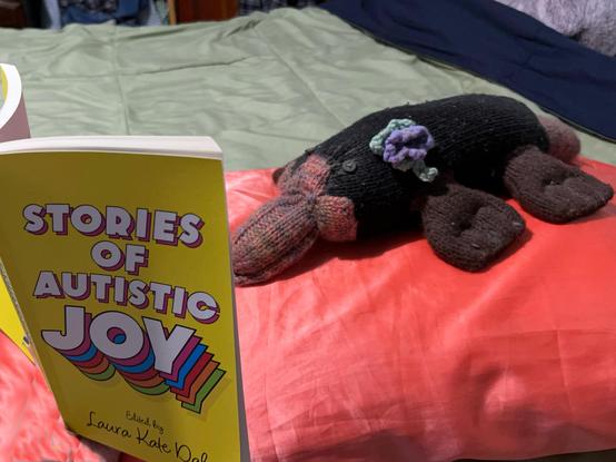An adorable handmade platypus stuffie rests on an orange satin pillow reading a book with a bright yellow cover titled Stories of Autistic Joy edited by Laura Kate Dale.