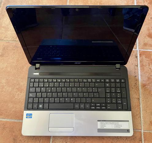 Imagen de un portátil Acer E1-571 abierto, mostrando la pantalla negra apagada y el teclado. El portátil es de color negro y plateado, con el logo de Acer visible en la tapa.  El teclado tiene una disposición estándar de QWERTY, y se ve un panel táctil debajo de él. En la parte inferior se puede ver una pegatina con especificaciones del modelo.
