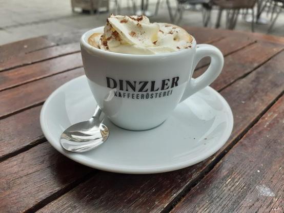 A cup of coffee with whipped cream on a saucer with a spoon, sitting on a wooden table.
