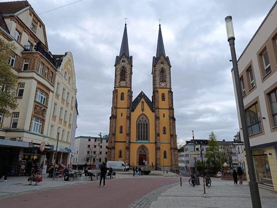 A church with twin spires stands in a town square on a cloudy day. Buildings flank the church, and people are visible on the plaza.