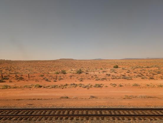 Another landscape photo featuring rail tracks at the bottom, endless pale blue skies above, and arid, orange-yellow desert in the middle.
