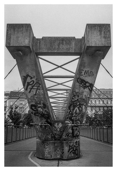 Black and white photograph of a pedestrian bridge (Siemens-Nixdorf-Steg over the Donaukanal, connects the 2nd and the 9th District) with a large, V-shaped concrete support structure in the center, covered in graffiti. Diagonal support cables cross behind the structure, leading the eye toward a row of classic European-style apartment buildings in the background.