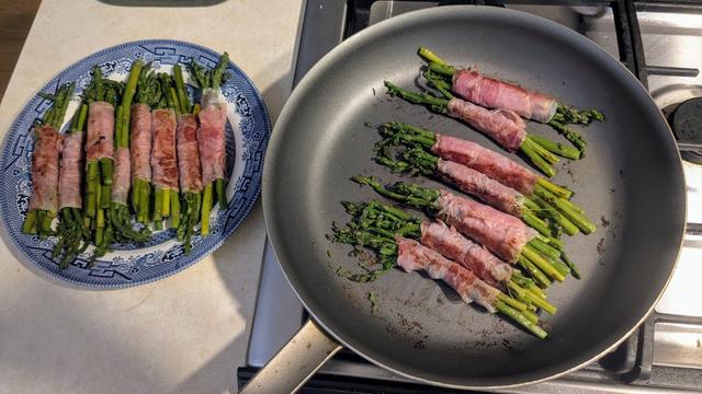 A blue and white plate and a large nonstick frying pan filled with stalks of green stems of asparagus wrapped in thin pink lunch meat.