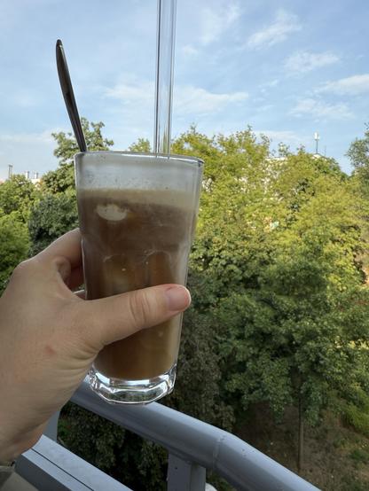 Glas mit Eiskaffee in einer Hand, im Hintergrund Bäume