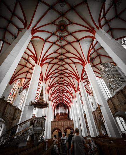 Innenansicht der Thomaskirche in Leipzig mit markantem rot-weißem Kreuzrippengewölbe, hohen weißen Säulen, historischer Kanzel und Blick auf die große Orgel im Altarraum. Besucher stehen im Kirchenschiff und bewundern die Architektur.