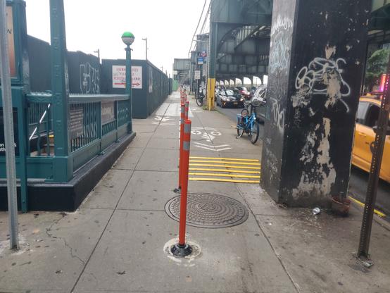 Sidewalk next to a roadway. On the left, a subway entrance. On the right, the roadway with large columns next to it (holding up an elevated subway). In the middle, the sidewalk is divided with delineator posts. The left side of the sidewalk is for marked for pedestrians, and the right side is marked for bikes (via sharrows, which is.. not what they fucking mean, NYC DOT assholes! Sharrows literally mean a shared lane BETWEEN CARS AND BIKES. The 2009 MUTCD states, "Shared Lane Markings shall not be used on shoulders or in designated bicycle lanes."). The right side bike area is marked in both directions, meaning two-way; and between the delineator posts and column, it's approx 3-4ft wide. For good measure they also added weird rumble strips. And of course the 3-4 width is almost entirely a metal manhole cover, which will be slippery in the rain.
