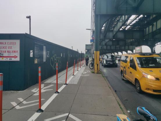 Further down the same sidewalk, viewed from the bike area. The left side no has a "sidewalk closed use pedestrian walkway" sign and some green walls, then the ped walkway, then the bike area, and then columns and then the roadway (with cars moving in it). The bikeway continues to narrow to 3-4ft wide, and there's a bike parked next to a column that further narrows the bikeway. Someone is biking just past it, to give you an idea of a 2.5ft wide cyclist just barely fitting (forget a cargo bike or trike.)
