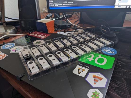 A box of pendrives tagged with different letters indicating the different distros, sitting atop a laptop covered in stickers.