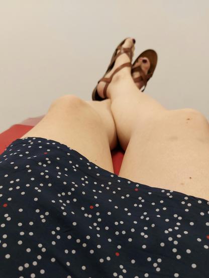 Man sieht einen blauen Rock mit Punkten und meine Beine und Füße mit Sandalen auf einer roten Liege