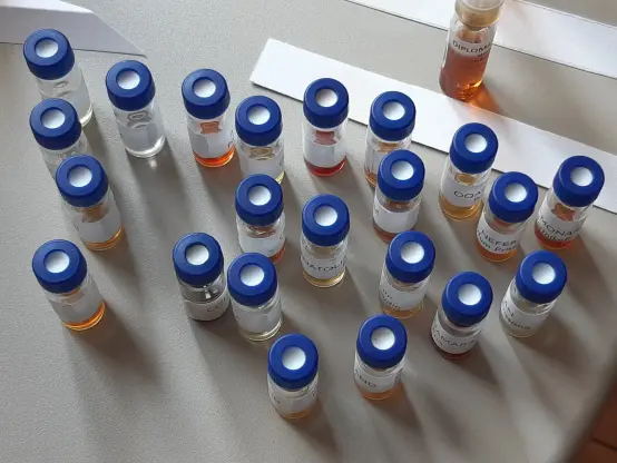 The image shows a collection of small glass vials arranged on a flat surface. Each vial has a blue cap and a label. The vials contain liquids of various colors, including clear, amber, and reddish hues. There are also several white strips of paper placed near the vials.