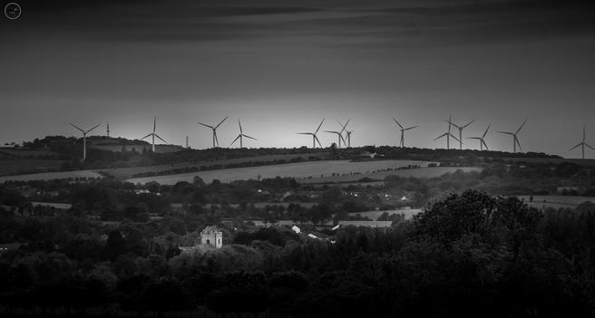 row of windturbines covering a hill side, heavy clouds above, woodland and partially obscured church in foreground