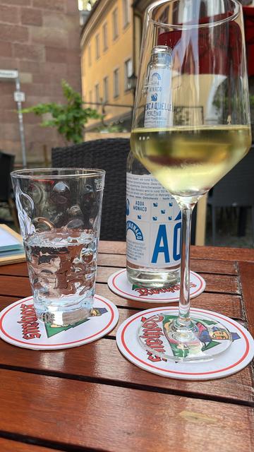 Ein Glas Weißwein und ein Glas Wasser auf einem Holztisch, mit einer Flasche Wasser im Hintergrund. Die Szene zeigt im Hintergrund verschwommene Gebäude und Grün, was auf eine Umgebung im Freien hindeutet.