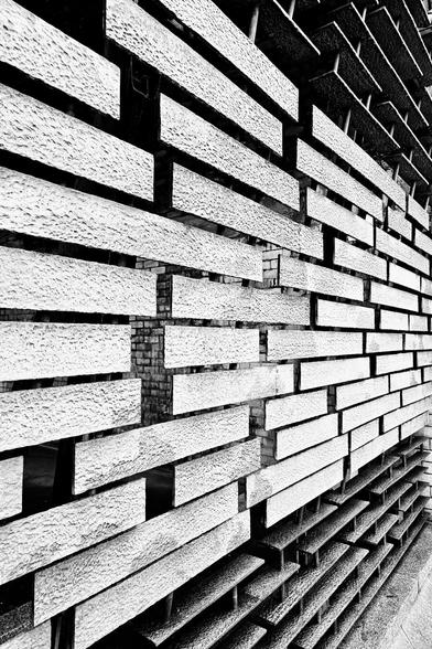 A high-contrast, black and white photograph of a textured wall, likely part of a modern building. The wall is composed of horizontally laid, rectangular blocks with a rough, uneven surface, creating deep shadows between them. The blocks are arranged in an offset, almost brick-like pattern, and the perspective looks upwards along the wall, emphasising the repetitive linear elements and the dramatic play of light and shadow.