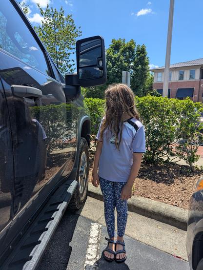 Photo of an 11 year old kid standing in a parking lot next to a dark gray "custom" Chevy Silverado. The truck appears to be stock (no after market lift kit). The kid's head barely reaches the bottom of the side view mirror and is lower than the bottom of the window.
