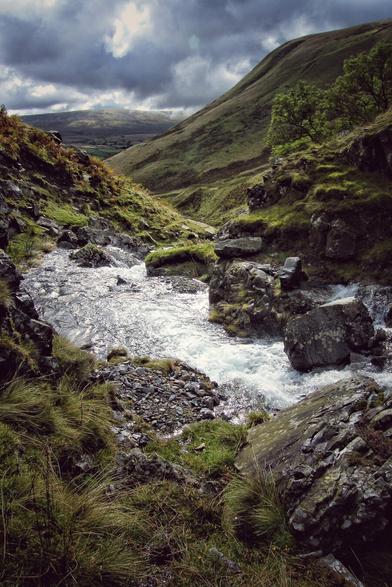 A dramatic view of a rocky stream cascading through a green valley near Cautley Spout in the Lake District. The water is bright and foamy, surrounded by dark, mossy rocks and lush green grass on the banks. Steep, grassy hills rise on either side under a moody, overcast sky with patches of bright light breaking through the clouds.