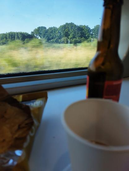 Un gobelet en carton blanc, une petite bouteille, un paquet de chips, sur fond de fenêtre du train donnant sur la campagne.