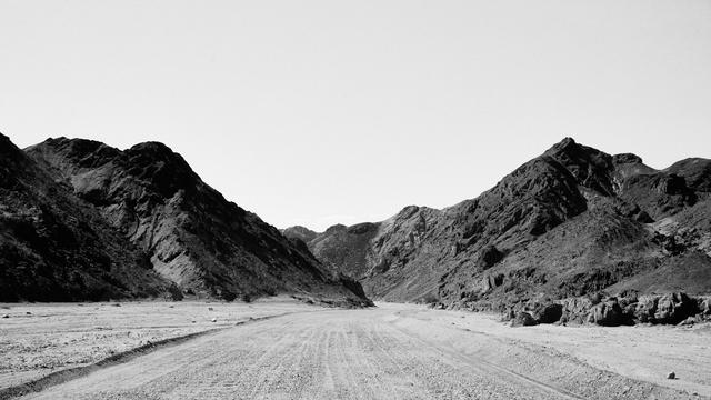 A wide landscape of a barren desert road leads through rugged, dark rocky hills under a clear sky. The terrain is dry and textured, with no vegetation visible.