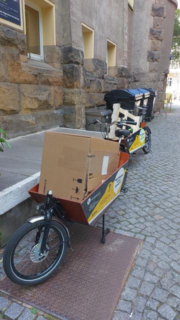 Zwei Lastenräder an der Laderampe der Poststelle der TU Dresden.  Das Ladefach des vorderen Rads ist mit einem großen Packet, das des hinteren mit einer Palette gefüllt.