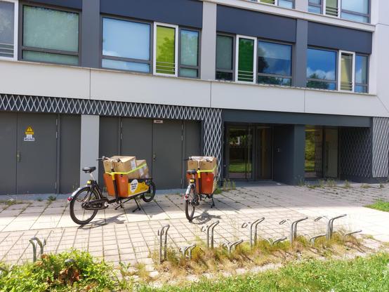 Zwei Lastenräder vor dem (Student:innen-)Wohnheim Gret-Palucca-Straße 11.  Beide sind mit vielen Kisten gefüllt, die mit einem Spanngurt am Rad gesichert sind.