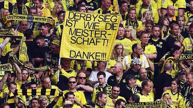 Ein dichtes Publikum in gelb-schwarzen Trikots und Schals bei einem Fußballspiel. Im Vordergrund hält ein Fan ein großes Banner mit der Aufschrift „DERBYSIEG IST WIE DIE MEISTERSCHAFT! DANKE!!!“. Die Menge zeigt Begeisterung und Unterstützung für ihr Team.