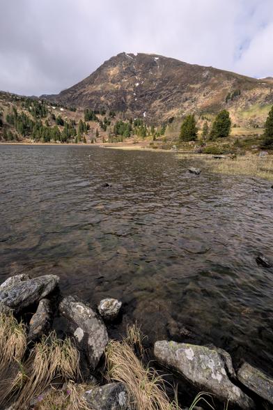 Vorne Steine und Gras vom Ufer, dann ein See und dahinter ein Berg. Der Himmel ist bewölkt.