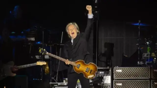Paul McCartney, en un concierto, toca un bajo Hofner violín de color marrón oscuro. Su brazo derecho está levantado en señal de triunfo. Lleva un traje negro y su pelo gris es visible. Se ve parte del equipo de sonido en el escenario detrás de él, incluyendo amplificadores y una batería parcialmente visible. Otro músico está parcialmente visible en el lado izquierdo del encuadre, tocando una guitarra.