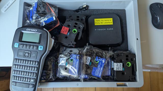 Ein Etikettiergerät und ein Labelprinter mit vielen Band-Kassetten in einem Euro-Box-Koffer.

Auf dem Labelprinter ist ein gelbes Label aufgeklebt:
"Hello, my Name is 
Printy McLabelface"