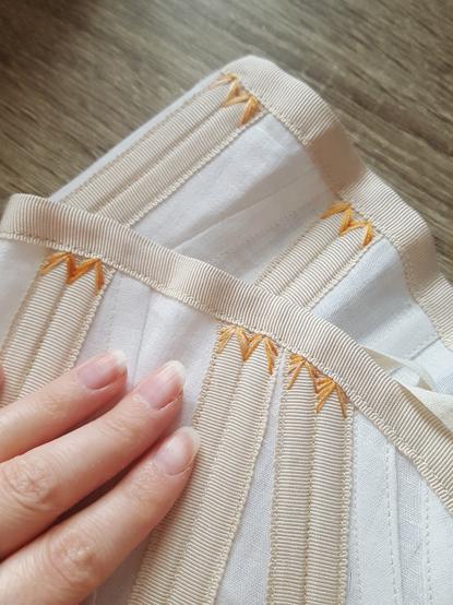 A close up photo of embroidery called flossing applied onto the ends of synthetic whalebone in their boning channels, made of petersham ribbon. The corset is white, the tape is a light beige, and the embroidery thread is variations of oranges, yellows and beiges. It stands out as a different hue, but not offensively. It's just a very simple V shape, with the legs around the bone and the tip of it securing the end of the bone. Sini's fingers in the frame.