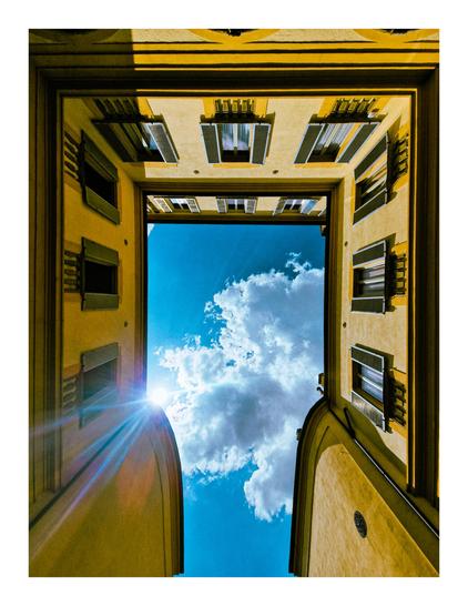 This photo is of a low-angle view looking up through a narrow, square opening formed by the walls of a building (Palazzo Albergati). The walls are a yellowish tone, with multiple windows visible on each side. Above, the sky is a vibrant blue with fluffy white clouds. Sunlight streams in from the bottom left, creating a lens flare.